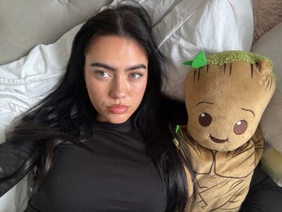 A woman with dark hair is lying on a bed with white sheets and a light gray quilted headboard. She is wearing a black long-sleeved shirt. Beside her is a stuffed Groot toy. The location is Aveley, United Kingdom.