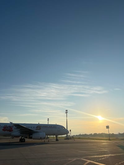 Here's a description of the image:

The photograph shows a Super Air Jet airplane on an airport tarmac under a sunny sky. The sky transitions from a deep blue on the left to a lighter hue with streaks of cirrus clouds near the sun. The sun is high in the sky, creating a glare.

The airplane is oriented horizontally in the frame, with its nose pointing toward the right. Details of the airplane, including its engines, landing gear, and fuselage markings (Super Air Jet, Say No to Drugs, Say Yes to Travel) are visible. Adjacent to the plane is an airport ground crew area, with yellow lined markings and runway edge lights. 

The location is Baji Mangngai, Indonesia.