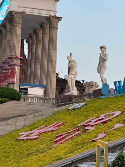 The image captures a street view in Nanshan District, China, featuring classical architectural elements, including a colonnade and statues of human figures and lions. A sign with red lettering is laid out on a green landscaped area. Modern elements, such as a waterfall feature and signage, intermix with the classical design. Banners attached to the columns have text in English and Mandarin that says "OF THE WORLD 世界之日". The image is rotated clockwise, giving a tilted perspective.