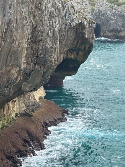 This image depicts a dramatic cliff face meeting the turquoise waters of the sea, likely in the coastal area of Llanes, Spain. The cliff is characterized by rugged, stratified rock formations in shades of grey and brown, with patches of green vegetation clinging to its upper reaches. The lower part of the cliff is covered in a darker, rougher material, possibly seaweed or barnacles, where the waves crash and foam. An overhang creates a shadowed area or a small cave-like opening. The sea is a vibrant blue-green, with white foam and ripples indicating wave action against the rocky shore and cliffs. The overall scene conveys a sense of natural grandeur and the power of the ocean.