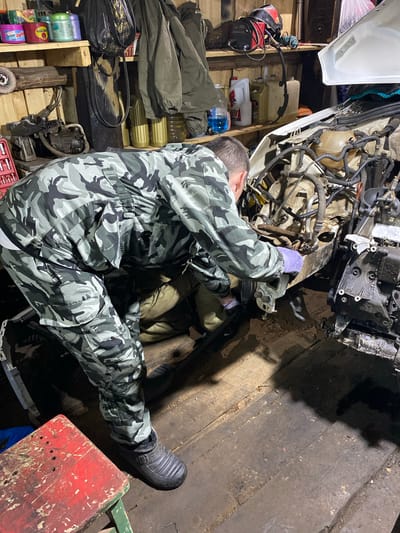 In a dimly lit garage, likely located in Kauchuk Stantsiya, Russia, a person wearing a camouflage jumpsuit is working on the engine of a white car. The car's hood is open, revealing a complex network of wires, hoses, and engine components. The person is bent over the engine, with one hand, covered in a purple glove, reaching into the engine bay. The garage appears cluttered, with shelves on the wall holding various items, including containers of liquids, tools, and what looks like a welding mask. The floor is made of wooden planks and is dusty and somewhat dirty, suggesting it's a working space. Another person is partially visible underneath the car, suggesting they are also involved in the repair. To the left of the scene, a red and green stool is positioned. The overall impression is that of a DIY auto repair session in a rustic garage environment.