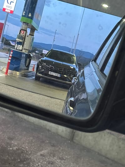 The image captures a scene at a gas station, likely in Kalugerovo, Bulgaria, as suggested by the context provided. The foreground is dominated by the reflection in a car's side mirror, showing the right side of the vehicle and the road. In the mid-ground, a black SUV is parked next to a blue gas pump. The SUV has its license plate visible, displaying the characters "KO E750 BO". The gas pump itself is tall and blue, with red and white striped posts nearby, and a red fire extinguisher mounted on its side. The background reveals a slightly hazy sky with scattered clouds and distant hills. The lighting suggests it is either dusk or early morning. There are no visible people in the frame.