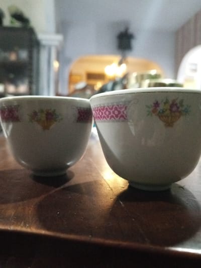 Two decorative ceramic bowls are positioned on a dark brown, polished wooden surface. The bowl on the left is smaller, featuring a central floral basket design with pink patterned sections on either side, and casts a distinct shadow. The bowl on the right is larger, displaying a prominent pink geometric pattern band near its rim and a central floral basket design, also casting a shadow. The wooden surface exhibits visible grain and minor surface imperfections. The background is out of focus, revealing an indoor environment in Longmiang, Laos, characterized by blurred architectural elements including arches, a light-colored pillar to the left, and indistinct furniture or shelving. Multiple blurred light sources are visible, illuminating the distant background. No people are present, and no actions or interactions are depicted beyond the static arrangement of the bowls.