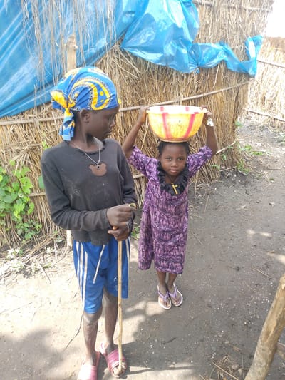 In Otulu, Nigeria, two young girls stand in front of a rustic dwelling constructed from thatch and covered with a blue tarp. The older girl, wearing a blue and yellow headscarf, a black long-sleeved shirt, and blue shorts with white stripes, holds a wooden stick. She looks to her right, her expression pensive. The younger girl, dressed in a purple patterned dress, balances a yellow and red bowl on her head. She wears a golden cross necklace. Her gaze is directed forward, her expression serious. The ground around them is dirt and debris.