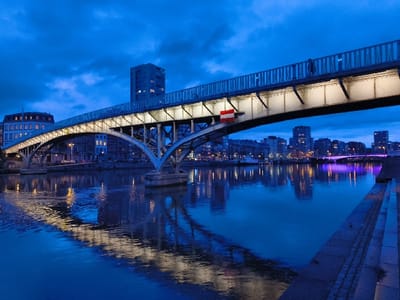 The image displays the Pont Albert I bridge over the Meuse River in Liège, Belgium, during a period of low light, likely dusk or night. The sky is a deep blue.
The primary subject is the multi-span metal arch-truss bridge, positioned centrally. Its structural components, including the arches and underside of the deck, are brightly illuminated by artificial lights, which cast distinct reflections on the calm, deep blue water of the river below. A pedestrian walkway with metal railings runs along the top of the bridge deck, and a single, indistinct human figure is visible walking on this walkway towards the right side of the frame. Attached to the underside of the bridge's framework, roughly in the middle-right section, is a rectangular sign featuring horizontal red and white stripes.
On the left bank of the river, a large, multi-story building with a rounded corner and numerous illuminated windows stands prominently. Further along this bank, additional urban buildings with illuminated windows are visible.
The immediate foreground on the right side of the image features a stepped stone or concrete embankment leading down to the river. Beyond the main bridge, on the right bank, a modern urban cityscape is visible, characterized by several tall buildings with illuminated windows forming a skyline. A second, more distant bridge further downriver is distinguishable by its distinct purple artificial lighting.