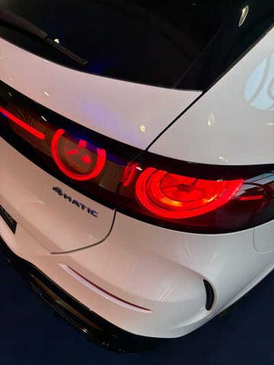 The media file is a close-up, angled shot of the rear of a white car with black accents. The car's red taillight is prominently featured, with its intricate internal design visible. The taillight has a circular element with smaller bulbs inside, and a vertical red strip below it. The word "4MATIC" is visible in silver lettering on the car's body, just below the taillight. The car appears to be in a showroom or a similar indoor setting, given the lighting and the dark, possibly blue, background which suggests a controlled environment. The lighting creates reflections on the car's glossy surface, highlighting its curves and the contrast between the white and black sections. There are no people visible in the media file. The image composition focuses on the design and details of the car's rear end, emphasizing its modern aesthetic. The lighting suggests it is either daytime with artificial lighting or nighttime. No specific weather conditions can be inferred. The location context provided is Parma, Italy, but there are no visual cues within the image itself to confirm this location.