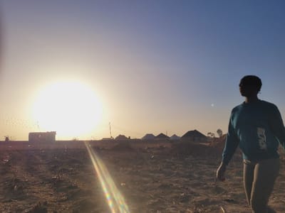 The scene, located in Shen, Nigeria, captures an outdoor environment at what appears to be sunrise or sunset. A single individual, largely silhouetted, walks from right to left across a dry, tilled field in the mid-ground. The individual wears a blue-green long-sleeved top with a "BLACK" inscription and light-colored trousers, with their head oriented towards the left. A large, low sun dominates the left side of the upper frame, generating a distinct lens flare diagonally across the lower-center. In the distant background, structures include a partially constructed or dilapidated multi-story building and several single-story, round-roofed dwellings. The sky is clear, transitioning from yellow-orange near the horizon to light blue overhead. Minimal vegetation is present on the arid ground.