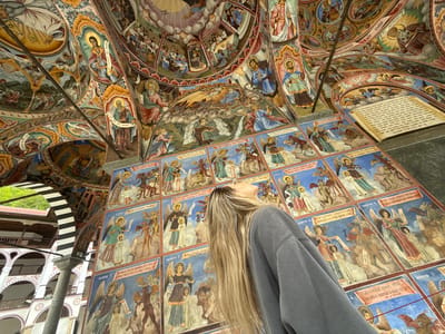 The image shows a woman with long blonde hair, wearing a gray sweatshirt, looking up at a wall covered in religious paintings. The paintings appear to be frescoes or murals depicting angels, demons, and biblical scenes. The wall is part of a structure with arched openings and striped columns, suggesting an indoor/outdoor space. The building appears to be richly decorated with religious frescoes on the walls and ceiling, including figures with halos and scenes from the Bible. The painting style appears to be Eastern Orthodox. The paintings are arranged in rectangular panels with text captions below. The image was taken at Rilski Manastir, Bulgaria.