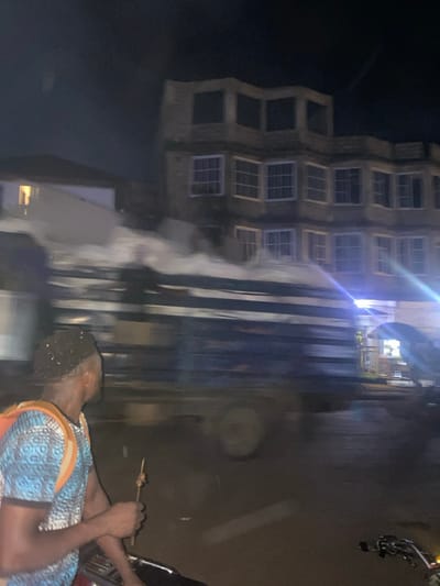 The image shows a man standing on the side of a road at night in Limbe, Cameroon. The man is wearing a blue and white patterned shirt and a backpack. He is holding a skewer with a piece of meat on it. He is standing next to what appears to be a motorcycle, which is partially obscured. A large truck, possibly a flatbed, is driving past him, blurred from motion, in the direction of a multi-story building with large windows. The truck is leaving a white trail behind it. The buildings in the background are lit and appear to be in a residential area. The road is wet. The scene is lit by streetlights. The coordinates provided place the location near the junction of Bonakanda Street and 1er Arrondissement in Limbe.