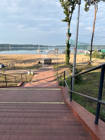The image shows an outdoor scene at a recreational area, likely a lakefront. In the foreground, a wide staircase with dark red brick treads and a yellow marking on the middle step leads downwards. A black metal railing runs along the right side of the stairs.
Beyond the stairs, a sandy beach extends towards a body of water. Several sailboats are docked on the water. Behind the beach, there are trees and some recreational structures, including what appears to be an adventure park with ropes and platforms suspended between trees. A tall metal pole with wires is also visible. The sky is overcast with scattered clouds. The scene appears to be located in Sława, Poland, a town known for its lake and recreational facilities. There are no people visible in the image.
