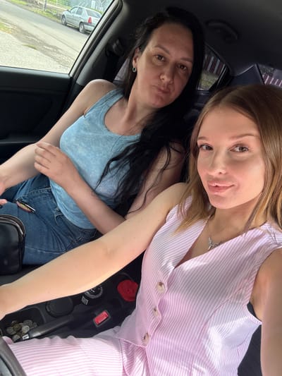 The image shows two women in the front seats of a car. The woman on the left has dark hair and is wearing a light blue tank top and blue jeans. The woman on the right has blonde hair and is wearing a pink and white striped jumpsuit with a silver necklace. They are both looking at the camera, and the woman on the right is smiling. The setting appears to be daytime, with sunlight coming into the car. The background outside the car shows a street and some buildings. The car interior shows a portion of the dashboard with some coins visible in a compartment. The image seems to be a selfie taken by the two women.