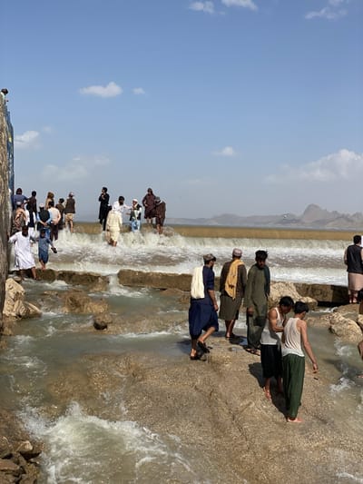 A daytime outdoor scene shows a group of people gathered around and in a body of water. The location is not Kim, Afghanistan. The water appears to be flowing over a series of low, rocky ledges or a constructed spillway. The water is murky and brown.

Several people are standing on the rocks, while others are wading in the water. The people are mostly men and boys, dressed in a mix of traditional and casual clothing. Some wear long robes and turbans, while others are in t-shirts and shorts.

In the background, a hazy mountain range is visible. The sky is blue with scattered clouds.

The overall impression is one of a leisure activity, with people enjoying the water and each other's company in a rural setting.