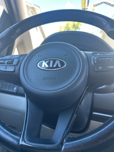 An exterior shot shows a black Kia steering wheel with the Kia logo visible in the center. The steering wheel has various control buttons on either side. The image appears to be taken from inside the vehicle, looking forward, with the exterior environment blurred in the background. It is daytime, and the sky is bright and clear. The scene is static, with no people or actions depicted. The image is not specific to Everett, United States of America.