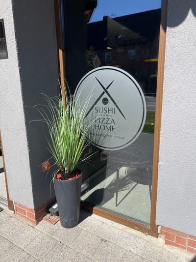 The image shows the exterior of a business with a large glass window. The window has a circular decal with the text "SUSHI AND PIZZA HOME" and a website address "sushipizzahome.pl". A tall, dark grey planter with a grassy plant is positioned in front of the window. A red and orange sticker with Polish text is visible on the building to the left of the planter. The building has a textured grey facade and a brick base. The ground is paved with a textured material. Reflections in the window show buildings and trees. This business is located in the city of Ustka, Poland.