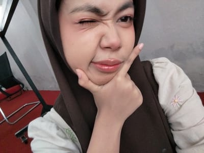 This is a close-up, waist-up photograph of a young woman wearing a brown hijab and a light-colored shirt with a floral embroidery detail. She is winking with her left eye and has her right hand resting on her chin, with her index finger pointing upwards. Her lips are slightly pursed and appear to have lip gloss. The background is dimly lit and appears to be an indoor setting, possibly a room with a red carpet and some furniture that is partially visible, including a chair and a metal stand. The overall mood seems playful and casual, as suggested by her expression and pose. The lighting appears to be artificial, and there is no visible text in the image.