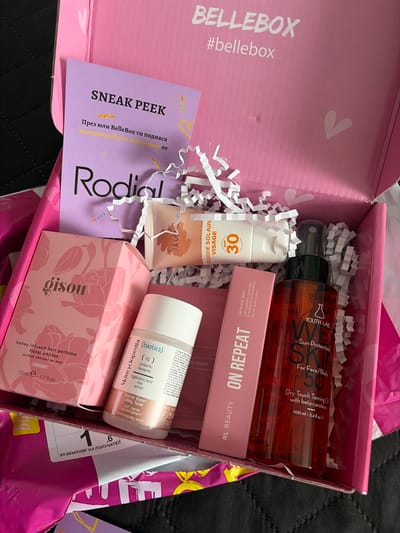 An overhead shot captures the contents of a pink "BELLEBOX" beauty subscription box, filled with various skincare and beauty products nestled among white shredded paper filler. The box itself is light pink with "BELLEBOX" and "#bellebox" printed in white on the side, and subtle white heart graphics on the interior.
The box appears to be freshly opened, suggesting an unboxing or product reveal. Products visible include a tall, amber-colored bottle of "YOUTH LAB Sun Protection SPF 50 Dry Touch Tanning Oil" (100 ml / 3.4 fl oz) for face and body. Below it is a pink boxed "NL BEAUTY ON REPEAT Lip Butter Balm" (10 ml / 0.34 oz) formulated with peptides, ceramides, and hyaluronic acid. A white bottle of "Skincyclopedia Barrier Boost Milky Toner" (50 ml / 1.69 fl oz) featuring probiotics, ferments, hyaluronic acid, rice, and ectoin, and stated as "Dermatologically tested," is also present.
Towards the bottom right, a pink box with a floral design holds "gisou honey infused hair perfume floral edition" (50ml e - 1.7 fl oz). A white tube with an orange cap for "FLUIDE SOLAIRE VISAGE SPF 30" (Sun Fluid for Face SPF 30), with a partially visible brand name that appears to be "Sk*v*," is placed to the left.
Two cards are visible: a purple "SNEAK PEEK" card with text in Bulgarian stating, "През юли BelleBox ти поднася вълнуващо бюти изкушение от Rodial" (In July BelleBox presents an exciting beauty treat from Rodial), displaying the "Rodial" logo. A white card with "ЕКЗЕМПЛЯР НА ПОЛУЧАТЕЛ" (Copy for Recipient) is partially visible on the right. The background is a dark, textured fabric. The scene is brightly lit. The location context provided is Novi Pazar, Bulgaria.
