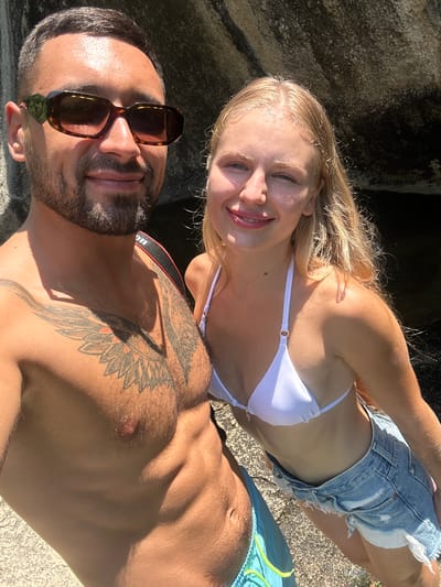 A man and a woman are taking a selfie outdoors. The man, on the left, has short dark hair, a beard, and is wearing sunglasses and blue swimming trunks. He has a tattoo on his chest and a camera strap around his neck. The woman, on the right, has long blonde hair and is wearing a white bikini top and denim shorts. They are standing near a rocky area with water visible in the background, possibly in Hoa Thang Ward, Vietnam. The sunlight is casting shadows on them.