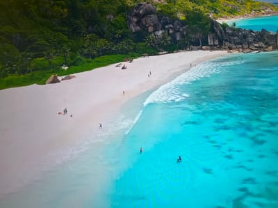 The image shows a tropical beach with white sand and turquoise water. A rocky outcrop borders the beach, covered in green vegetation. Several people are on the beach and in the water, some swimming and others walking along the shoreline. The location is not in Basingstoke and Deane, United Kingdom; it is a tropical beach.