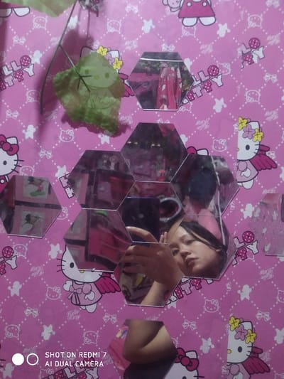 A vibrant pink Hello Kitty wallpaper serves as a backdrop for a selfie reflected in a collection of hexagonal mirrors. The reflection shows a person holding a phone, capturing their face and arm. A whimsical touch is added with a green leaf bearing a Hello Kitty design, positioned near the top left corner. The image also contains the text "SHOT ON REDMI 7 AI DUAL CAMERA" at the bottom left corner.