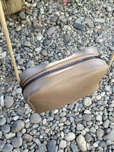 A light brown rectangular shoulder bag, appearing to be crafted from synthetic leather, is suspended above a densely packed surface of grey, brown, and white pebbles. The bag features a black zipper with a beige tape running along its top, secured by a silver-toned pull tab positioned on the right side. A thin, braided light beige strap extends upwards from both the left and right sides of the bag, exiting the frame. Visible stitching defines the bag's edges. A blurry dark vertical object is partially visible in the upper left background. This scene is located in the city of Panton, Indonesia.