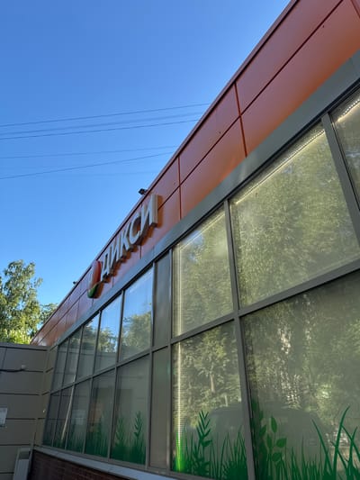 The image shows the exterior of a "ДIКСИ" supermarket in Bykovo, Russia. The building features an orange facade with a large glass window display. The "ДIКСИ" logo, a red apple with white lettering, is prominently displayed on the orange panel above the windows. The windows themselves are decorated with a green grass pattern at the bottom and are illuminated by a strip of white LED lights running along the top edge. Reflections of trees and foliage are visible in the glass. The sky is a clear, bright blue, with some power lines visible in the upper portion of the image. The image is taken from a low angle, looking up at the building.