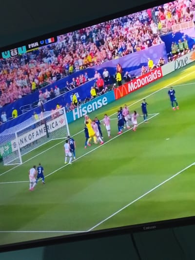 This image is a photograph of a soccer match being broadcast on a television screen. The screen displays a live view of the game, with players on a green field and spectators in the stands. In the upper left corner of the screen, there is text that appears to be part of the broadcast overlay, including "FIFA" and what looks like country flags, likely indicating the teams playing. Sponsorship logos are visible along the side of the field, including "McDonald's" and "Hisense." A goal net is in the left portion of the frame, with players in blue and white jerseys near it. The overall scene is a typical broadcast of a professional soccer game. There is no indication of the location being Tolú, Colombia, within the image itself.