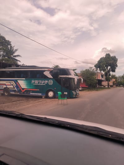 The image depicts a static scene in Reuhattuha, Indonesia, viewed from inside a vehicle, with the dashboard and windshield wipers partially visible in the foreground.
The primary subject is a large, double-decker passenger bus, colored teal and white with blue and red stripe accents. It is parked on a dirt shoulder beside an asphalt road. The bus prominently displays "PM 702 #" on its side, along with the number "05" near its front wheel. Smaller text, partially visible below "PM 702 #", appears to read "PUTRA NAGA ATJA PERMAI". A bright green plastic chair is positioned in front of the bus, near the road's edge.
The scene is set under a cloudy sky, with soft, diffused light suggesting either early morning or late afternoon. To the left of the bus, dense tropical foliage, including palm trees, is visible. To the right, along the road, more trees and several buildings, including a red-bricked structure, line the street. A sign on one of the distant buildings in the background might read "KECAP NAGA" or a similar phrase, though it is not entirely clear. No people are visible in the image. The overall atmosphere is quiet.