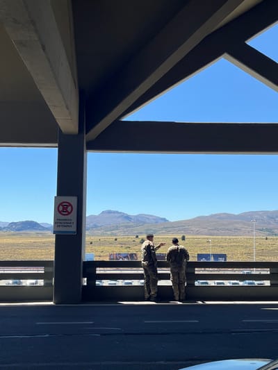 Two soldiers in uniform stand on a platform, possibly a parking garage, with a mountain range and open field visible in the background. A "no parking" sign is attached to a pillar, written in Spanish, "Prohibido Estacionary Detenerse." A structure with beams frames part of the scene, contrasting with the open sky above.