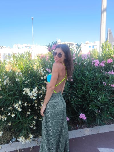 A woman stands outdoors in front of a flowering bush, facing away from the camera and looking over her shoulder. She has long, dark hair and is wearing sunglasses, a bright blue bikini top with neon yellow straps, and a long, green, snakeskin-patterned skirt. The background includes white buildings, a lamppost, and a bright blue sky. The scene appears to be located in the city of Paphos Municipality, Cyprus.