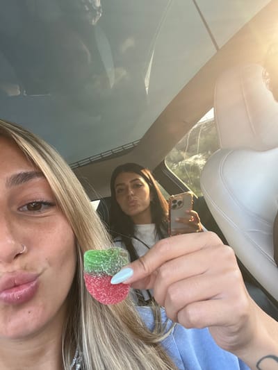 The image depicts two women and a partial view of a third individual inside a vehicle, presumably in Rosh Haayin, Israel. In the foreground, a blonde-haired woman, visible from the forehead to the chin, poses with slightly pursed lips. She has a silver nose ring on her left nostril and holds a heart-shaped, sugar-coated gummy candy, half green and half red, in her right hand, which has light blue fingernail polish. In the background, seated to the right, a dark-haired woman, visible from the chest up, also poses with pursed lips. She is holding a gold-colored smartphone in her left hand. A small portion of a third person with dark skin is visible on the far right, obscured by a white headrest. The vehicle features a panoramic glass roof, reflecting clouds and displaying a view of rolling hills with sparse vegetation and a metal guardrail in the distance. Bright sunlight enters the vehicle from the right side.
