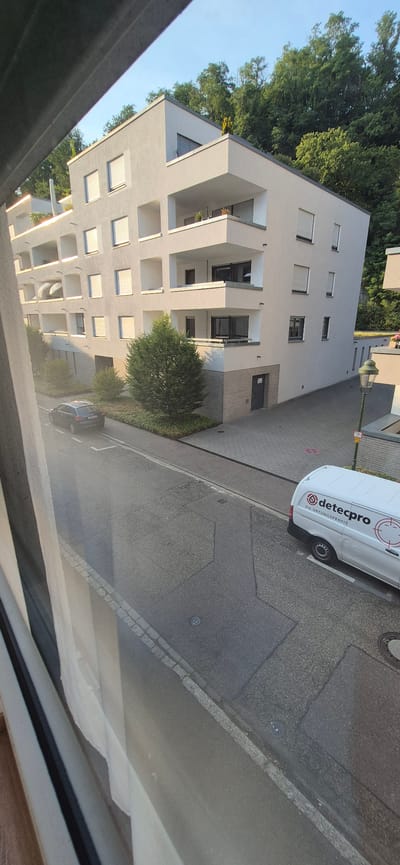 This media file depicts an exterior view of a modern apartment building with a tree-lined background. The building has a light-colored facade with numerous balconies and windows. A street runs alongside the building, with a paved sidewalk and a grey asphalt road.
Several objects are visible in the scene. A white van with the logo "detecpro" and the text "DIE ORTUNGSProfis" is parked on the street. A dark-colored car is also parked on the side of the road. A street lamp stands on the sidewalk, and there are several manhole covers on the road. Green trees and bushes surround the building and line the street. The sky is clear and blue, suggesting fair weather. The sunlight casts shadows, indicating it is likely daytime, possibly late afternoon or early morning given the angle of the light.
The scene appears to be a residential area in Baden-Baden, Germany, likely a quiet street in front of an apartment complex. There are no people visible in the frame. The overall impression is one of an ordinary day in a suburban or urban setting. The building's modern architecture, the presence of parked vehicles, and the street infrastructure are key details. The text on the van is clearly legible.