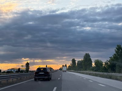 The image depicts a highway scene at dusk, with a black Kia Sorento in the foreground driving on the right lane. The car's license plate reads "CB 6879 EO." A white truck is visible further ahead in the same lane. The sky is filled with dramatic, cloudy formations, with pockets of orange and yellow light from the setting sun breaking through. On the left side of the highway, there are industrial buildings and streetlights, suggesting a commercial or industrial area adjacent to the road. The right side of the highway is bordered by a metal guardrail and a line of trees. The location is identified as the highway near Tsaratsovo, Bulgaria.