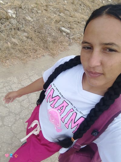A woman is standing outdoors on a path or road in El Cercado, Venezuela. She has dark brown hair in two long braids and brown eyes. She is wearing a white t-shirt with "MIAMI" in pink letters and a pink design, as well as pink pants with a white drawstring. She has a maroon backpack slung over her shoulder. The background consists of dry vegetation and a dirt path. The image appears to be a selfie.