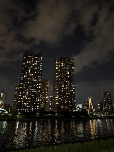 The photograph captures a nighttime cityscape in Koto, Japan, likely from a vantage point across a body of water. Dominating the scene are several tall, modern skyscrapers, their facades illuminated by numerous lit windows. The buildings appear to be residential or office complexes, with a dense pattern of lights suggesting multiple occupied floors.

In the mid-ground, a distinctive bridge structure with a unique, arching design stands out, illuminated against the night sky. This bridge's architecture is a prominent focal point. To the right and further back, another skyscraper with a red light at its apex is visible.

The foreground features a body of water, likely a river or bay, reflecting the city lights in shimmering patterns on its surface. The water is dark, indicating the depth and time of day. Along the bank of the water, a grassy area and some low-lying vegetation are visible, with a metal fence or railing running along the edge. The sky is dark and filled with scattered clouds, some of which are catching the city's ambient light. The overall impression is one of urban activity at night, with the interplay of light and shadow on the architecture and water creating a dramatic effect.