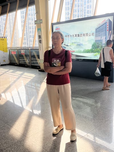 The image depicts an elderly Asian woman standing with her arms crossed in front of a large digital billboard. She is wearing a maroon t-shirt and light-colored pants. The billboard behind her displays an advertisement for "中铁十九局集团有限公司" (China Railway 19th Bureau Group Co., Ltd.), featuring a rendering of a modern building and a high-speed train. To the right of the woman, a man in a white tank top and black shorts is walking away, carrying a plastic bag.

The scene appears to be an indoor public space, possibly a transportation hub or a commercial building, given the presence of the billboard and the architectural elements visible. Sunlight streams in from a high angle, creating strong diagonal shadows on the polished floor, suggesting large windows or a skylight. Signs with Chinese characters are visible on a pillar to the left, one of which reads "跳磴" (Tiaodeng), indicating the location as Tiaodeng, China. Another sign shows a directional arrow and the number "18". The overall environment is modern and clean, with a focus on architectural lines and reflections.