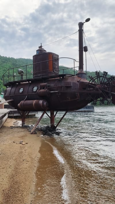 An abandoned, rusty, steampunk-style submarine sits on the sandy shore next to a body of water in Nikola, Russia. The submarine is elevated by several metal supports and appears to be made of riveted metal. It has multiple portholes, a large smokestack, and what appears to be a paddle wheel at the stern. A wooden pier extends from the left side of the frame, leading towards the submarine. The water is calm with small waves lapping at the shore, and the background features a tree-covered hillside under a cloudy sky. The overall scene depicts a static, weathered structure in a natural, waterfront setting.