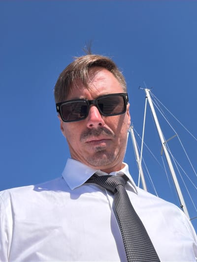 A low-angle, close-up selfie of a Caucasian man wearing a white collared shirt and a dark-patterned tie is captured against a clear blue sky. He has short brown hair, some of which is sticking up, and is wearing dark sunglasses. A faint mustache and goatee are visible. Behind him, the white masts and rigging of two sailboats are prominent against the sky. The man appears to be standing on a boat, as indicated by the visible rigging and masts. The lighting suggests it is a sunny day. The location is the Village of Sag Harbor, United States of America.