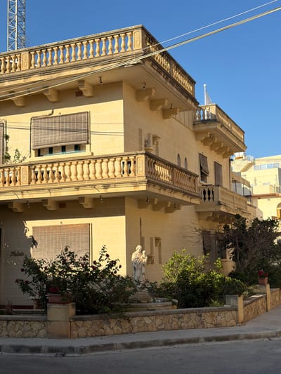 The image depicts a sunlit, corner view of a pale yellow, two-story building in Xagħra, Malta. The building features ornate balconies with balustrades and decorative stone elements. On the lower level, a statue of what appears to be the Virgin Mary holding the infant Jesus stands prominently in a small garden area. The garden contains rose bushes with red blooms and other greenery, enclosed by a low stone wall. The street in front of the building is paved, with a sidewalk bordering the garden. In the background, other buildings, including one with an external staircase, are visible under a clear blue sky. A tall, metal tower structure is also visible in the upper left corner of the frame. The lighting suggests it is daytime, likely late afternoon, given the long shadows. The building has blinds on some of its windows. A nameplate reading "Madonna" is visible on the left side of the building at the ground level.