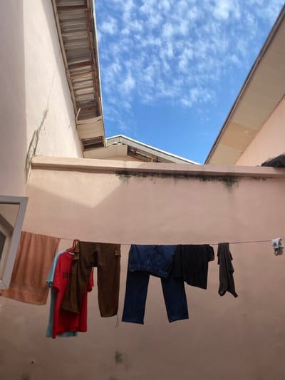 The image shows a clothesline with various garments hanging in a narrow outdoor space between two buildings. The buildings are painted a pale pink color, and their roofs are visible at the top of the frame, with corrugated metal roofing on one side and lighter colored material on the other. The sky is blue with scattered white clouds. On the clothesline, from left to right, are an orange towel, a light blue t-shirt, a red t-shirt with "GO" written on it, a pair of brown pants, a pair of dark blue jeans, a black t-shirt, a dark grey shirt, and a pair of white and green socks clipped to the line. The scene appears to be in a residential area, possibly a courtyard or alleyway, with the details of the buildings suggesting an urban environment in Dumnga, Nigeria.