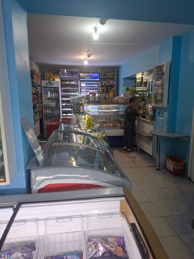 The image shows the interior of a small convenience store in Jitiqara, Kazakhstan. The store is brightly lit with fluorescent ceiling lights. The walls are painted a light blue.
In the foreground, there is a chest freezer with its lid open, revealing white wire baskets containing packaged frozen goods. Partially visible on the right is another freezer or display case, with a glass lid and a red and silver exterior.
Behind these freezers, a display of refrigerated goods is visible, likely drinks or dairy products, with clear glass doors. Further back, a boy wearing a dark, patterned shirt and dark pants stands at a counter, looking at something in his hand. To his right, shelves are stocked with various products, including bottled drinks and snacks.
The store appears to be well-stocked with merchandise arranged on shelves and in refrigerated display units. The flooring is tiled in a light color. There is a trash can with a red base and a yellow liner visible on the right side of the image, near the counter. A small round table with metal legs is attached to the wall on the right. The overall impression is of a functional, everyday retail environment.