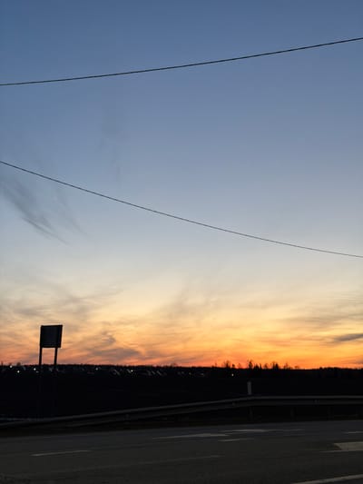 The image shows a sunset scene near a road in Kuregovo, Russia. Overhead power lines cross the pale blue and orange sky. A dark treeline and faint lights are visible in the distance, along with a road sign. The edge of a road runs along the right side of the frame.