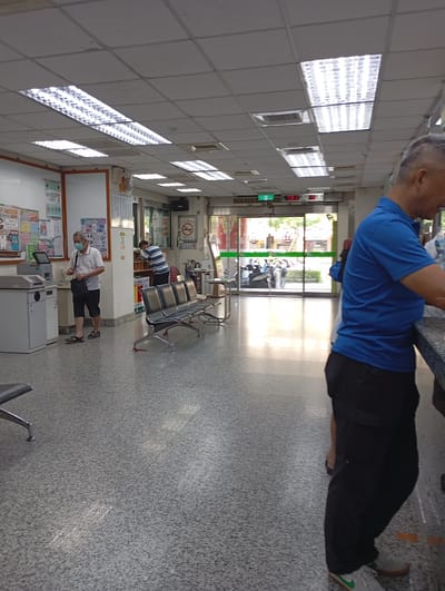 The image depicts the interior of what appears to be a clinic or hospital waiting area in Kaohsiung, Taiwan. Several individuals are present: one man wearing a mask and carrying a black bag is interacting with a payment kiosk. Another man in a striped shirt is standing by a counter. A man in a blue shirt and black pants is standing at a counter near the camera. There are rows of metal benches for seating. The room is well-lit by fluorescent ceiling lights. Signage is visible in Traditional Chinese. The exterior is visible through a glass entrance; motorbikes are parked outside.