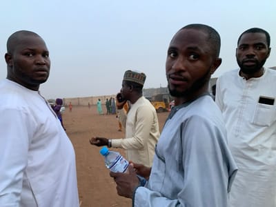 The image shows four men in Sokoto, Nigeria, standing in a dusty, open-air area. The man in the center of the image, wearing a light blue robe, is holding a clear plastic bottle of water in his right hand. The man to his left is wearing a white robe. He is facing the camera and holding a cell phone in his left hand. The man to the right of the center man is wearing a white robe and has a smartphone in his left shirt pocket. The man furthest to the right, also wearing a white robe, is looking towards the camera. Behind these men, a dirt road extends into the distance. There are other men walking in the distance, some wearing traditional clothes, some in modern clothing. There are several cars parked in the distance, as well as a light-colored pickup truck. The sky is cloudy and appears to be dusk.