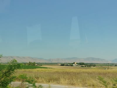 The image depicts a rural landscape under a clear, pale blue sky. In the foreground, dry, tall grass is visible along a paved road. Beyond the road, a vast field of green crops extends towards a cluster of buildings and trees in the midground. Further in the distance, rolling hills or mountains form the horizon. A thin reflection of what appears to be text is visible on the upper left portion of the image. The scene suggests a sunny day in a cultivated agricultural area. The location is identified as the Municipal Unit of Larissa, Greece.