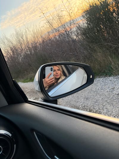 The image depicts a blonde female capturing a selfie reflected in the driver's side exterior mirror of a vehicle, positioned in Vrba, Montenegro. The perspective is from inside the car, looking out. The lower left of the frame shows the dark, textured interior panel of the car door. The white exterior mirror reflects the woman, who holds a light-colored smartphone in her left hand, aimed at the mirror. She has light-colored hair, a faint smile, and wears a light-toned garment. Beyond the vehicle, a gravel road or shoulder is visible, leading to a dense line of bare or sparsely leaved bushes and small trees. The sky above displays warm hues of orange and pink, characteristic of either sunrise or sunset.