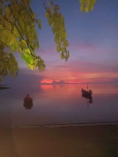 The image depicts a serene sunset scene at Baan Wok Tum, Thailand. The sky transitions from deep blue to pink and orange hues near the horizon, reflected in the calm water. Two boats are present: one close to the shore appearing as a dark mass and a second, further out, featuring an elevated element like a mast or antenna. A sandy beach borders the water. A branch with green leaves hangs from the upper left corner, framing the scene.