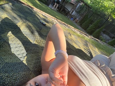 The image depicts a young woman lying on a patterned mat in a garden. She is lying on her side, with her right arm bent at the elbow and her hand resting near her face. She is wearing light-colored clothing and several bracelets on her wrist. The mat has a geometric pattern in dark green and yellow. Sunlight casts shadows across the mat. In the background, there is a grassy area, a wooden fence, several evergreen bushes, and part of a building with a tiled roof. The scene suggests a relaxed outdoor setting, likely in a backyard in Galabovo, Bulgaria, given the presence of Mediterranean-style vegetation and architecture.