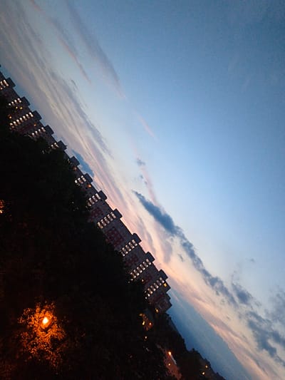 The image depicts a tilted perspective of apartment buildings at dusk. The sky is a gradient of light blue, transitioning to hues of pink and orange near the horizon, with scattered clouds. The buildings, with multiple stories and visible lit windows, are lined up diagonally from the bottom left to the top right of the frame. In the foreground, a bright, orange-yellow street lamp illuminates dark foliage. The scene appears to be an urban residential area in Most, Czechia.