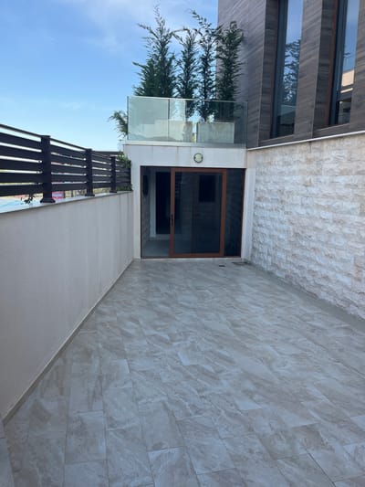 This image depicts an outdoor patio area with modern architecture. The patio is paved with large, light-colored, marble-patterned tiles. To the left, a light-colored textured wall runs along the edge of the patio, topped by a dark slatted fence. Beyond the fence, a portion of a swimming pool is visible. The background features a section of a building with a stone facade and large, dark-framed windows with a vertical wooden accent design. On the upper level, a glass railing encloses a terrace area with several tall, slender trees. In the center of the image, a sliding glass door with a wooden frame leads into the building. The sky above is a clear blue with a few wisps of clouds. The scene suggests a contemporary residential or commercial property in a well-maintained outdoor setting.