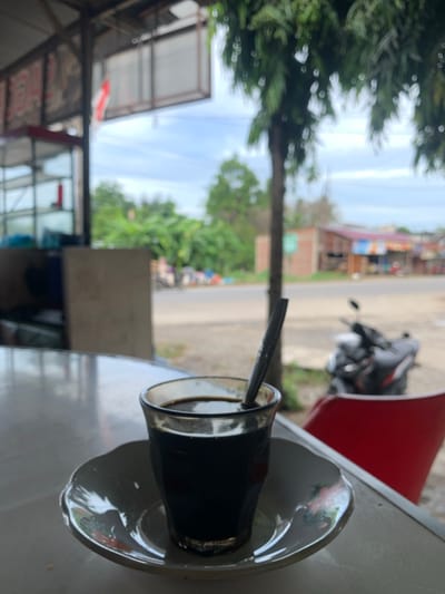 The image depicts a glass of dark liquid, presumably coffee, on a saucer. The glass is placed on a reflective surface, likely a table, in the foreground. A dark stirring stick is partially submerged in the liquid. In the background, an outdoor scene unfolds. To the left, a structure with shelves and possibly merchandise is visible, suggesting a shop or stall. A red and white flag is also seen hanging. Beyond this, a road runs horizontally, with a motorcycle parked on its right side. Buildings line the road, and lush green trees are scattered throughout the landscape. The sky above is partly cloudy. The scene is consistent with a roadside eatery or shop in a tropical environment, potentially in a city like Meuse, Indonesia, given the presence of tropical vegetation and typical Southeast Asian architecture. The overall atmosphere is casual and relaxed.